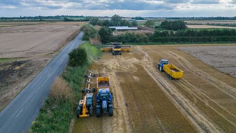 Photo of farm machinery working a wheat field along side a road cutting through the landscape. Likly Drone footage